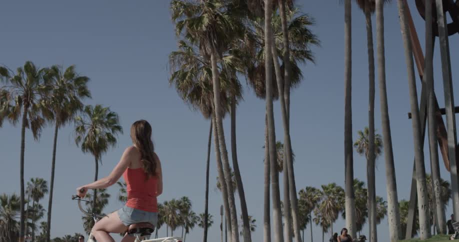 A woman rides a bicycle past tall palm trees