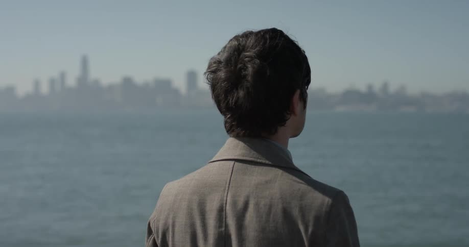 A man is standing with short hair and a jacket overlooking a large large lake or ocean with a city on the horizon on a bright sunny day