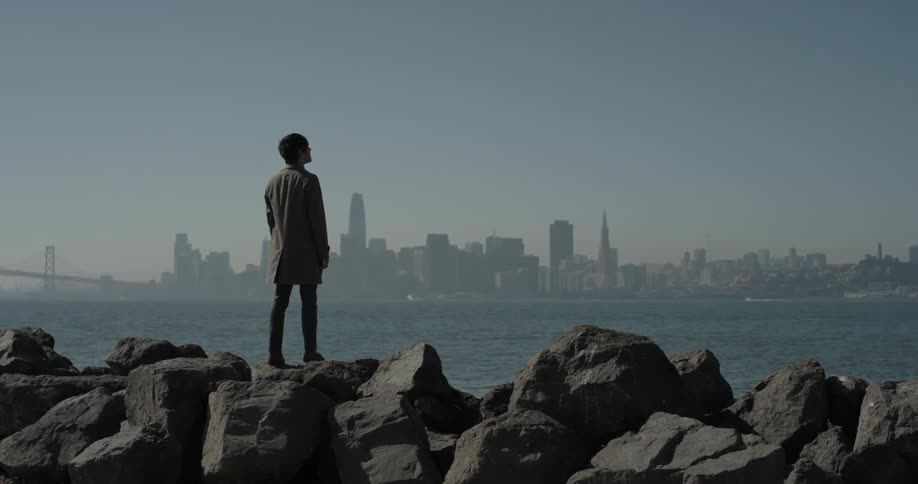 Man stands on large rocks looking out at a body of water and a city
