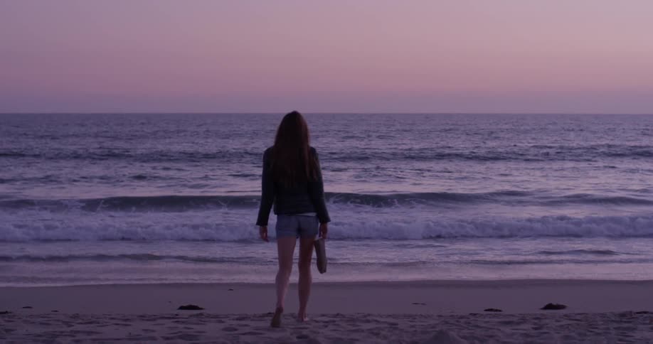 A woman in a jacket walking towards the ocean carrying her shoes in her hand