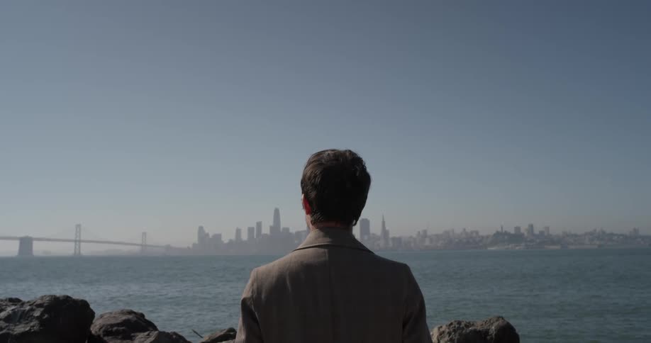 A man stands by the rocks of an ocean or great lake overlooking a large city off in the distance of a bright sunny day