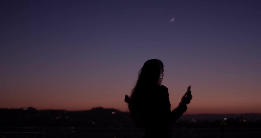 Woman waves her arms while holding a phone at sunset