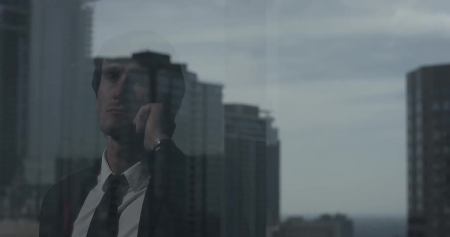 A man can be seen through a window wearing a suit with the window reflecting the city he's looking at