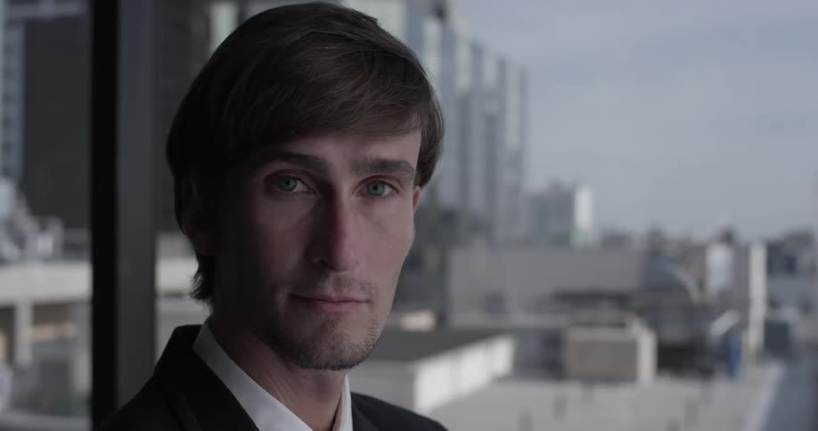 A man with short hair stares out the window and then turns to face the camera and blinks a few times