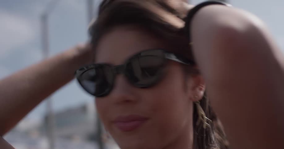 A woman wearing a cutoff shirt puts on sunglasses and dances in a car with the hood off and the wind blowing through her hair as she grabs her head