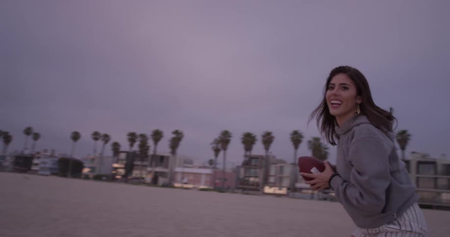 A woman wearing a sweater is playing catching or playing football at the beach on a cloudy sunset day at the beach with palm trees and building lights