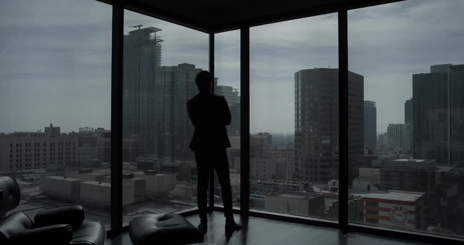 A man stands in the corner of a room with several large windows overlooking the cityscape