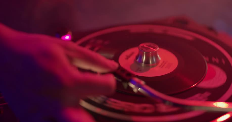 A dj puts a new vinyl record onto the mixer and adjusts it to the part he wants on stage of a red and blue lit concert