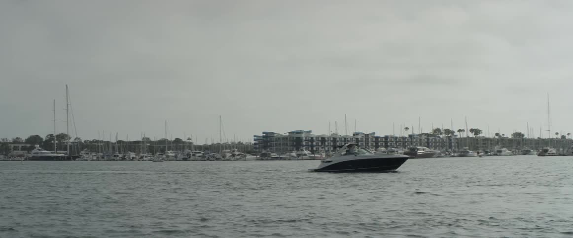 A ski boat moves across the water with some buildings and a marina in the background