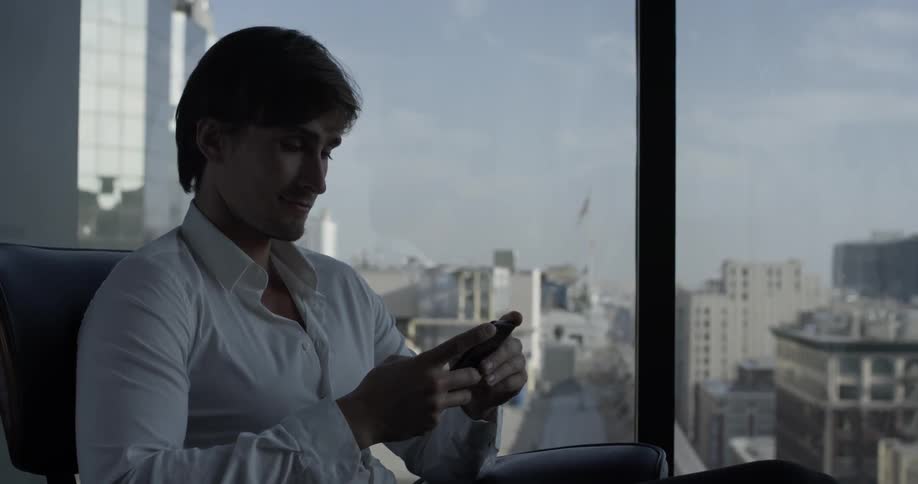 A man in a white shirt with short hair sits down in an office chair in an office looking at his phone in his hands