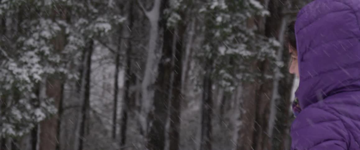A woman wearing a purple coat is walking and smiling in a wooded area as snow falls
