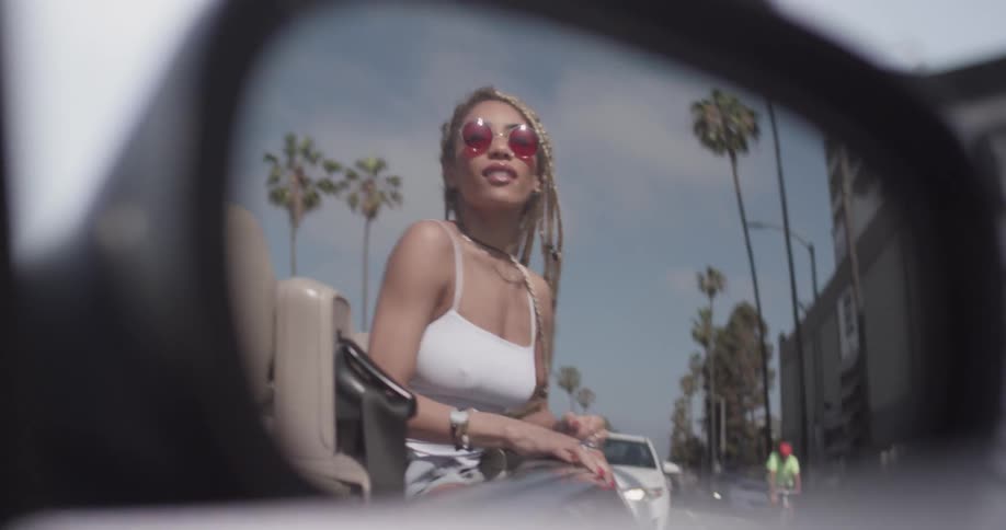 A side mirror of a convertible reveals a woman dancing in the back seat