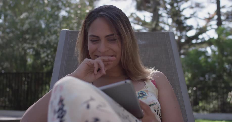 A woman sits on a pool chair as she reads on a tablet and smiles