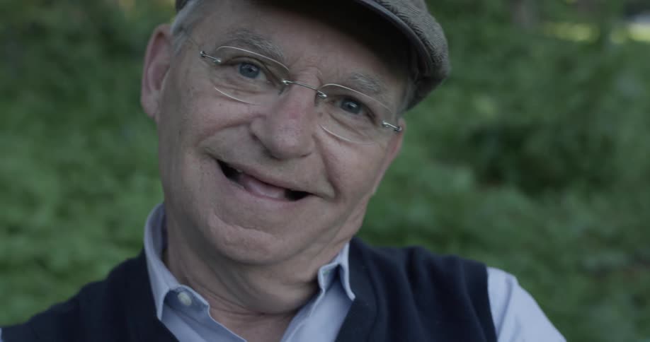 A man looks at the camera and has a laugh with glasses a hat and vest on in a park on a sunny day
