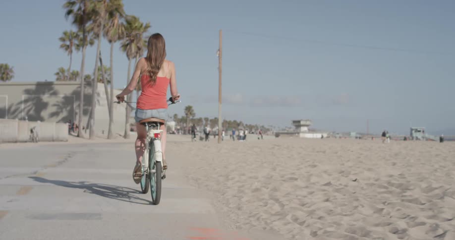 Women ins short clothing and a tank top with long beautiful hair rides her bike down the sidewalk at the beach near the ocean