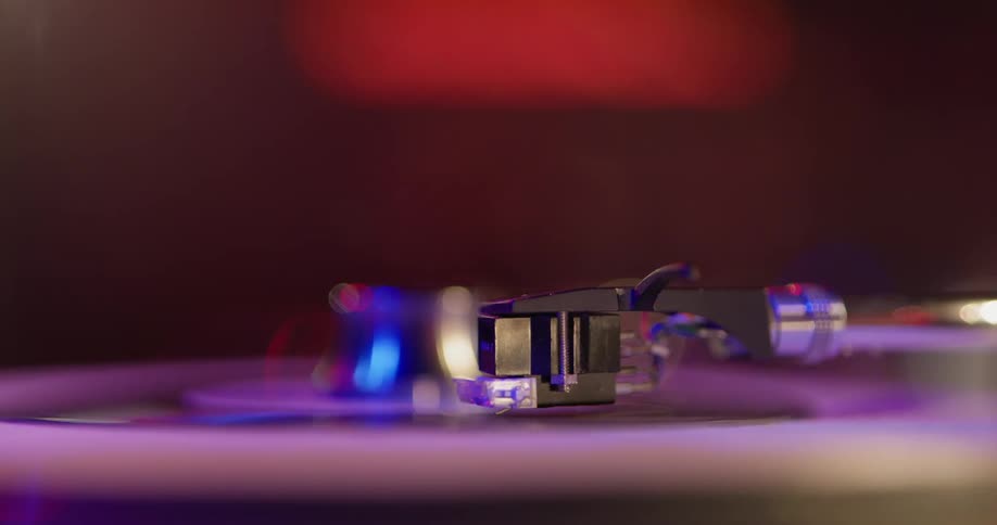 A record player needle is shown close up in detail while a record spins underneath with red and blue lights shining above