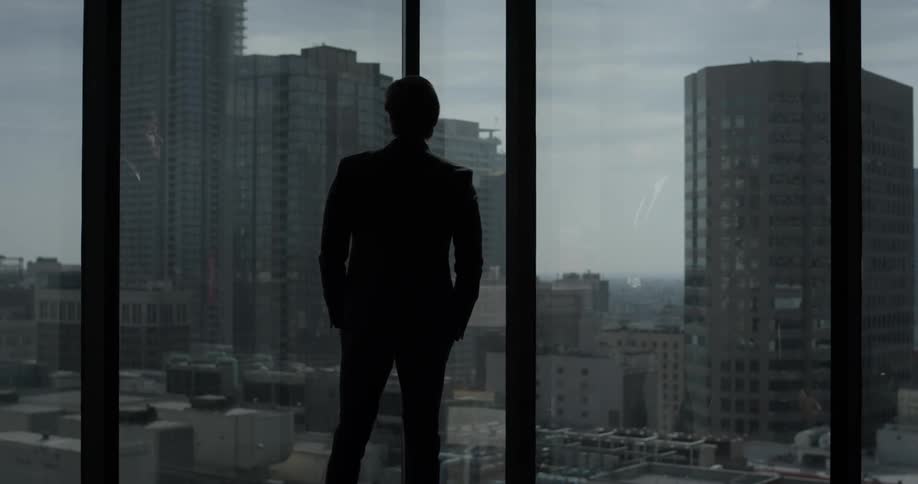 A man walks over to large windows and looks at the city