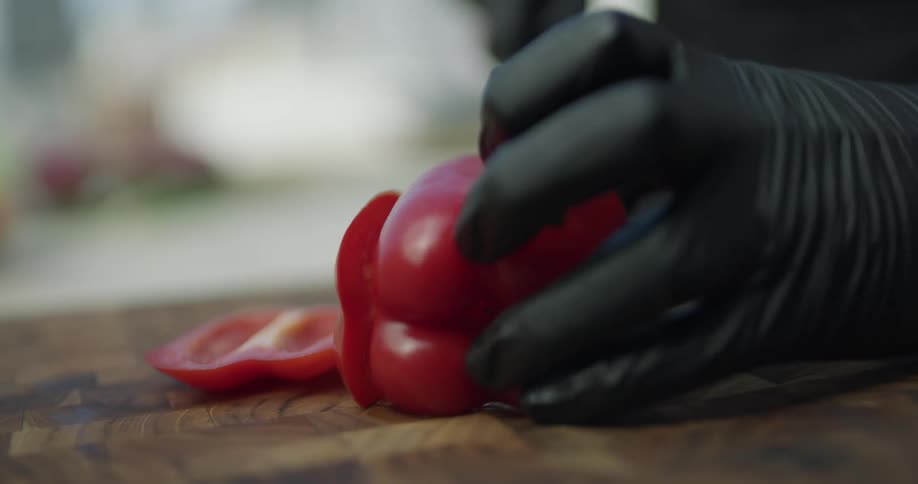 A chef wearing gloves chops a red bell pepper on a brown wood cutting board while wearing black gloves