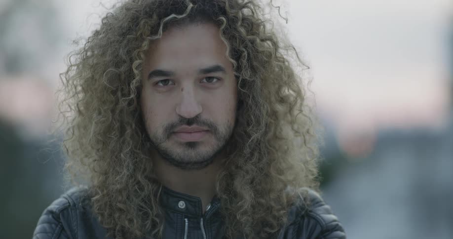 A man opens his eyes and stands there with long hair and light facial hair on a dimly lit day