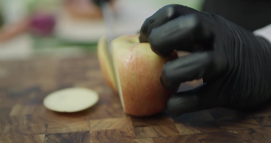 A chef wearing black gloves cuts an apple into slices on a checkered wooden cutting board