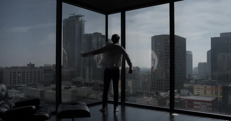 Man walks to the corner of an office room and throws on a white shirt there buttoning it up and staring out a window at the city