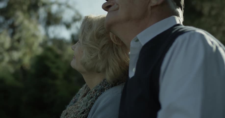 A elderly women and a man sit together on a park bench and smile with one another as he puts his arm around her