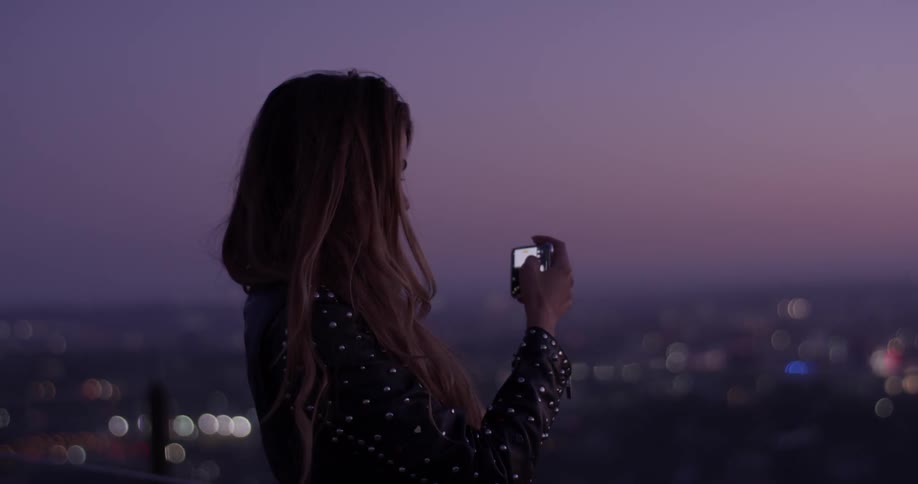 A woman stands up on a hilltop overlooking the city below holding her camera taking a picture after the sun has set and the city lights dot the landscape