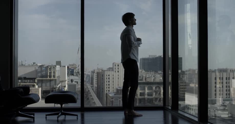 A man puts on a shirt in the corner of an office room with large windows overlooking the city
