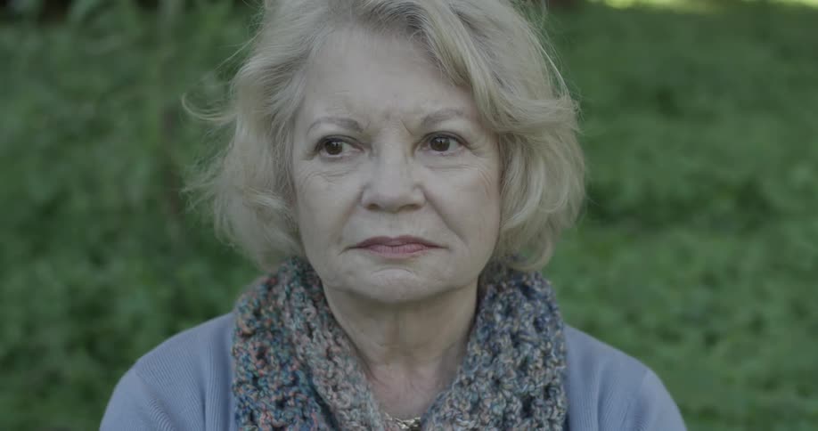 An elderly women with blond hair sits at a park and moves her head around looking at things