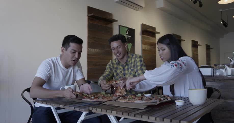 Three people sit discuss and laugh as they talk about something serious and eat pizza together on a table at the office with coffee mugs