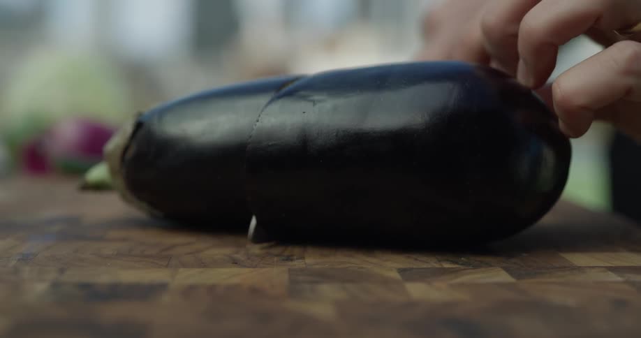 Someone chops a knife into an eggplant on a wooden cutting board then slices it in two