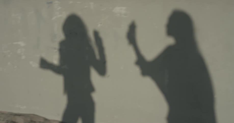 The shadow of two women can be seen on a cement wall as they walk on the sidewalk holding ice cream and chatting with one another