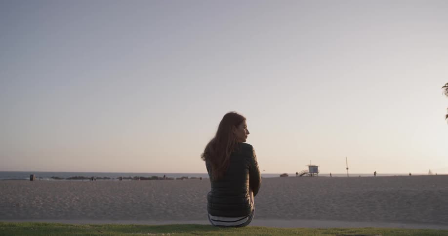 Woman sits on a sidewalk next to a sandy beach at sunset