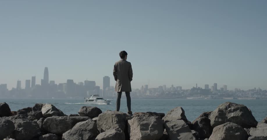 Man stands on rocks and watches boat in the ocean in front of him