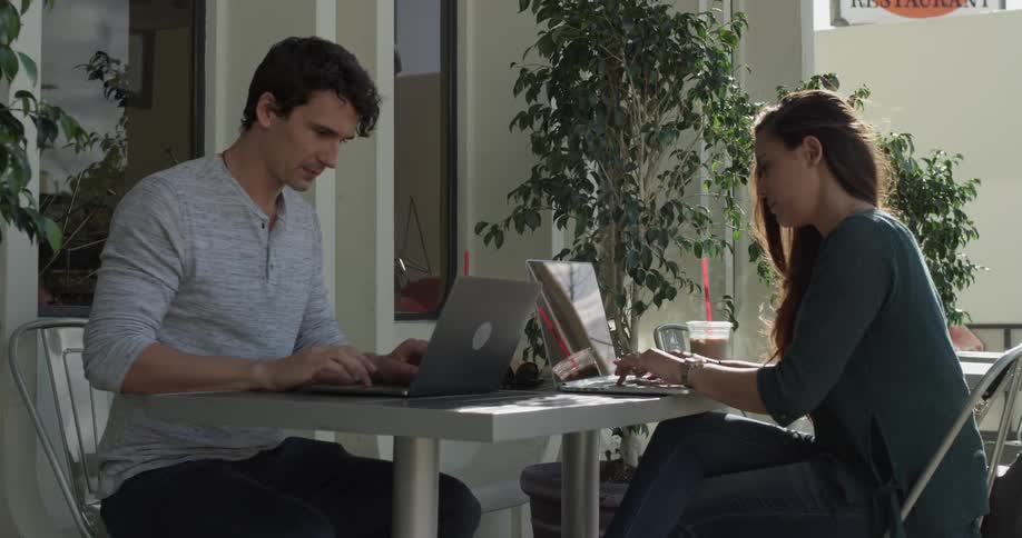 A man and a woman sit at a desk working on laptops and talking