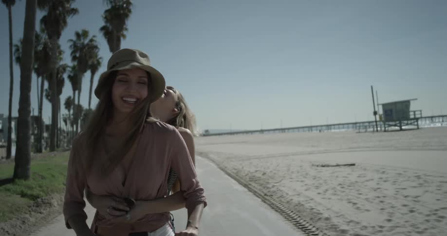 Two women ride a scooter together by the beach