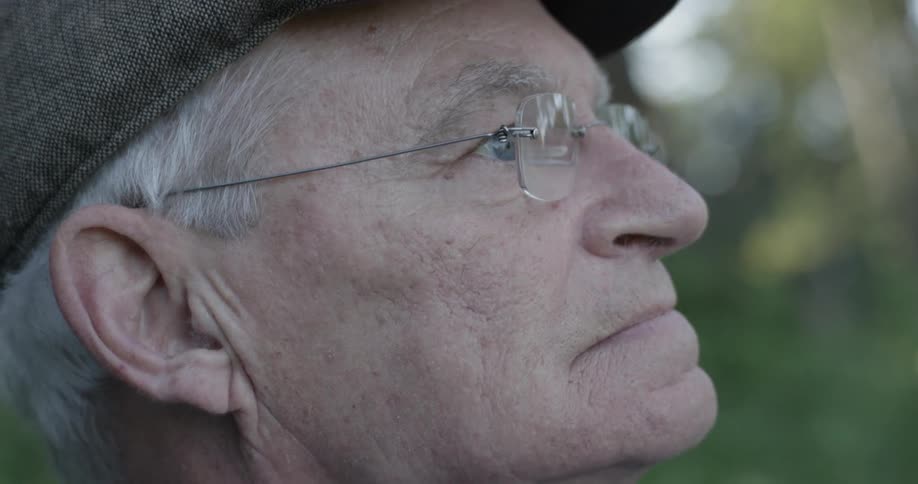 Old man with gray hair wearing glasses and a hat looks off into the distance at a park on a sunny day