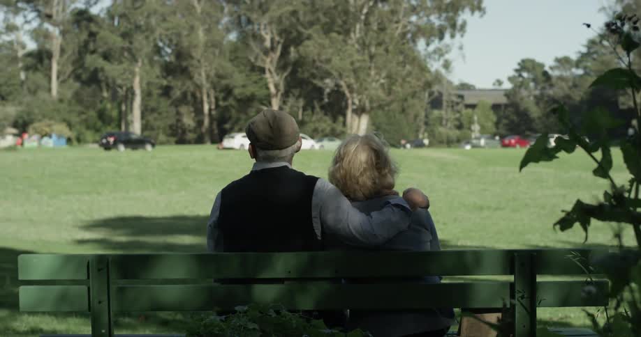 Man puts arm around elderly women on bench and touches soothingly while leaning head on shoulder almost and overlooking a sunny park