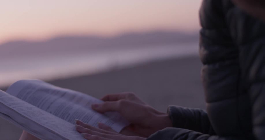 A woman sits on a beach reading a book during a sunset