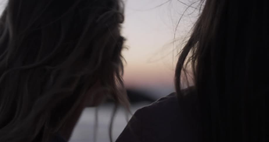 Two women talk to each other and watch the ocean at dusk