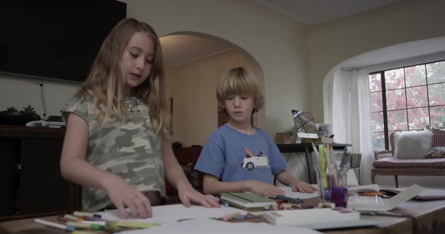 Two children fold papers while standing at a table indoors