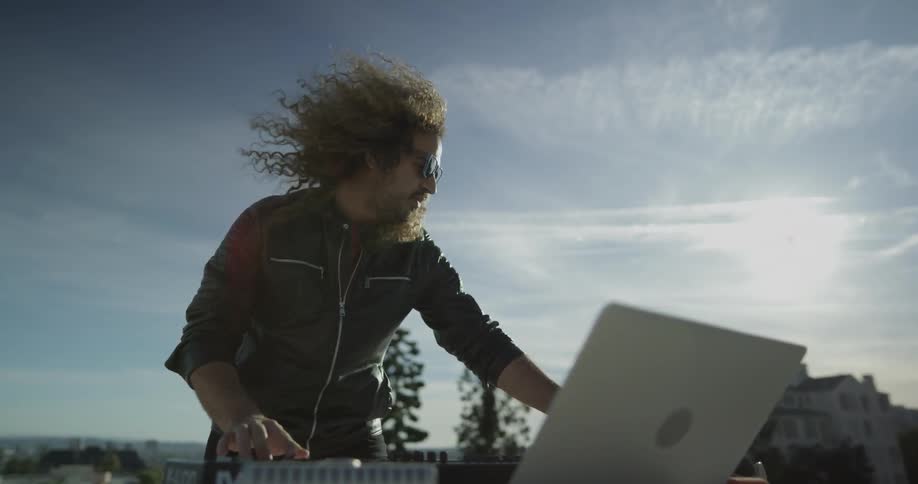 A man with long curly hair dances as he plays his keyboard in the setting sun