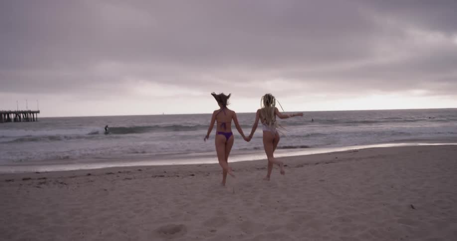 Two women hold hands as they skip across the sand toward the ocean