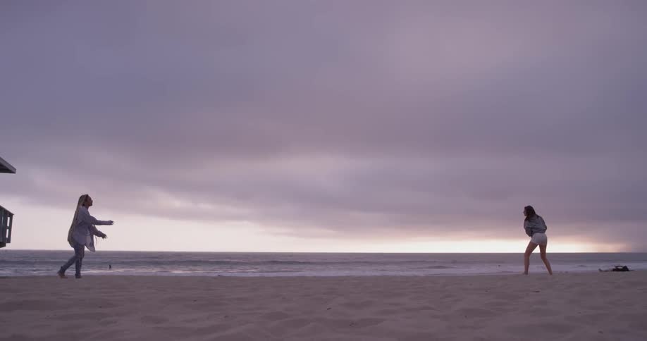 Two women play catch football on the beach of the ocean on a cloudy sun set day