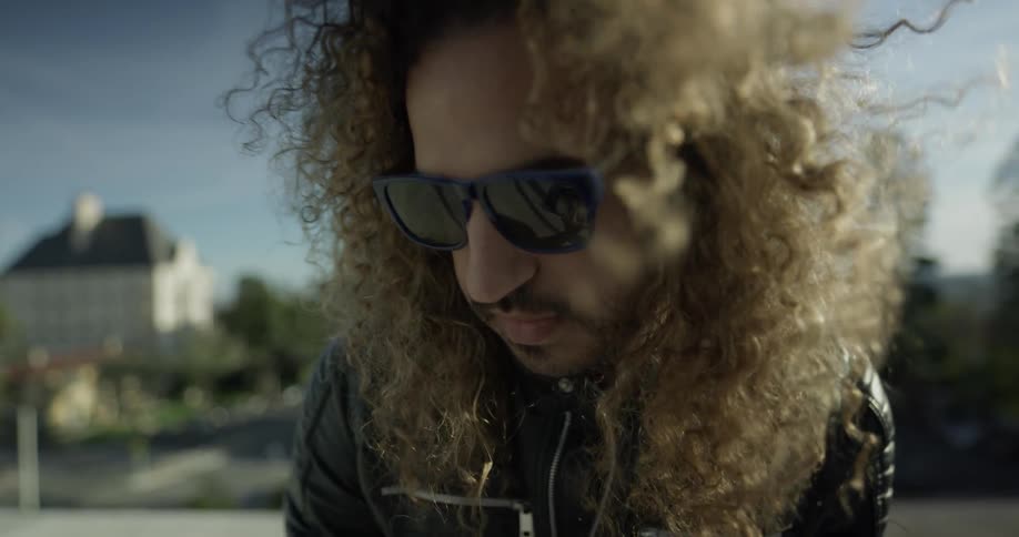 A mean with long hair wearing sunglasses plays a keyboard outside