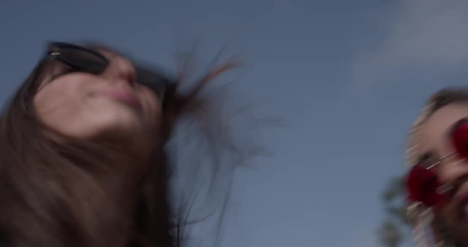 Two women dance in a car with the roof top off and the wind blows heavy on their waving hair wearing sunglasses and tank tops