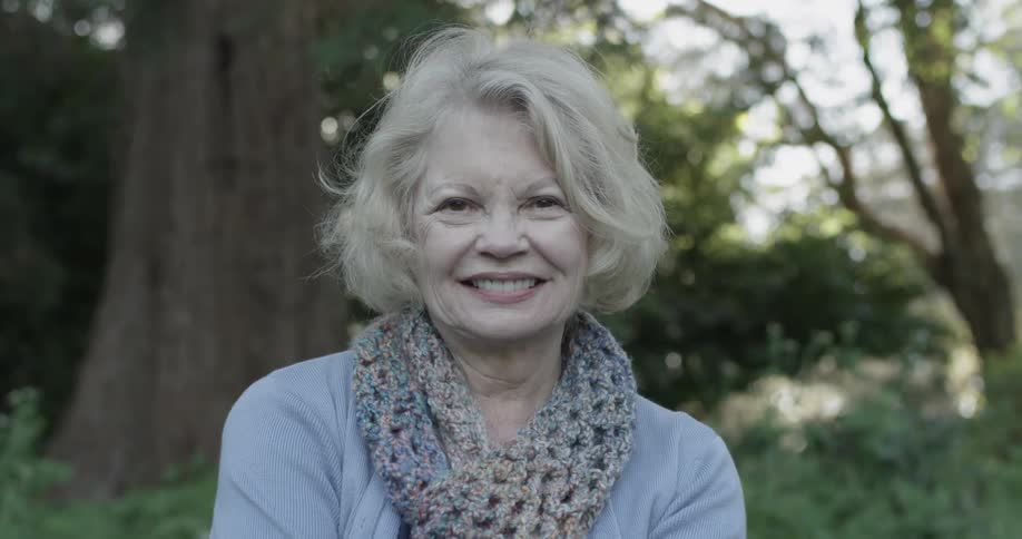 An elderly women looks at the camera and smiles while wearing a blue shirt and scarf on a bench she sits on a sunny day at the park