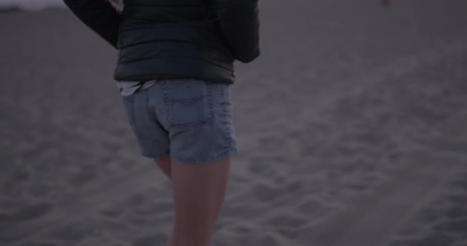A woman in jean shorts and a coat walks on a beach