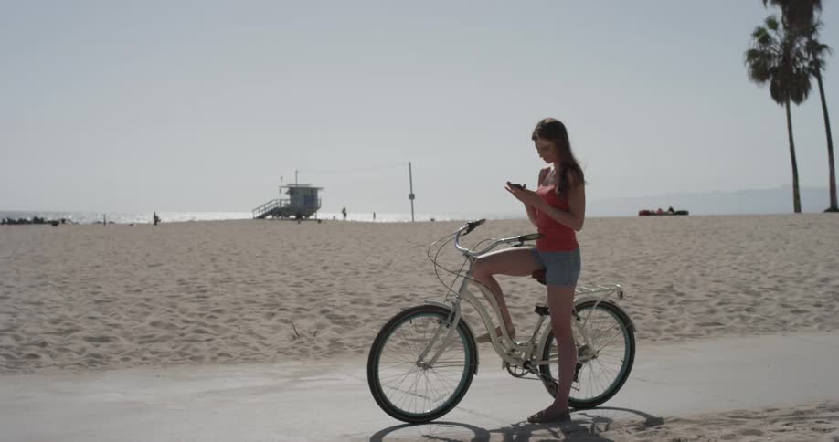 Woman sitting on a bike on the sidewalk by the beach looking at her phone