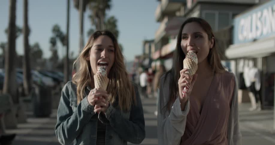 Two women with ice cream walk down the sidewalk happily pointing things out and dancing